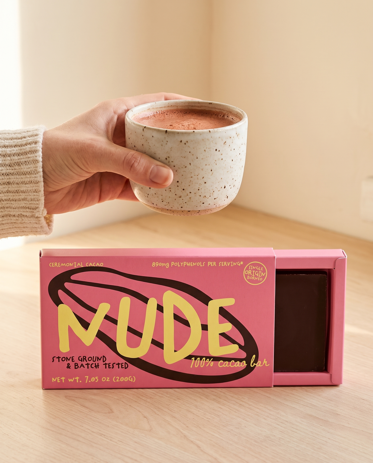 NUDE sleeve half slid open showing the 100% cacao bar, a cup of frothy drinking cacao lifted above