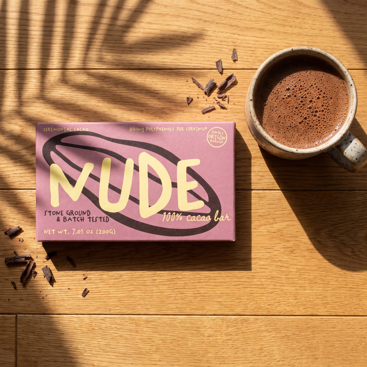 A pink NUDE cacao box on warm oak wood beside a speckled ceramic cup of frothy drinking cacao, with palm-frond shadows and cacao shavings
