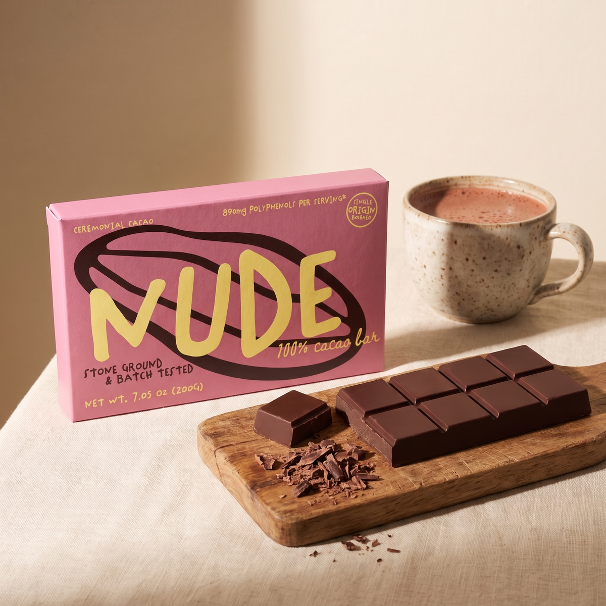 The pink NUDE cacao box behind its dark ten-piece scored cacao block on a wooden board, one piece cut off beside rough shavings, with a speckled cup of frothy drinking cacao