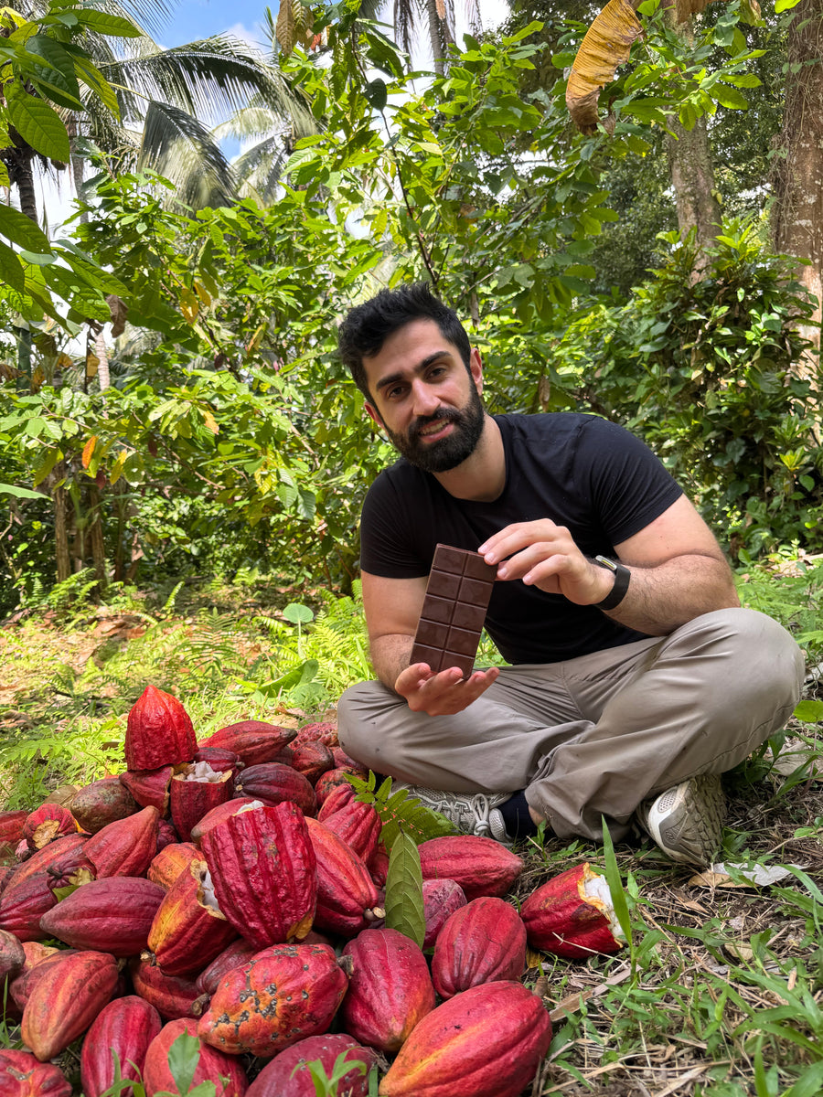 Ali, NUDE's founder, sitting in a cacao grove holding the bare ten-piece bar among a pile of red pods