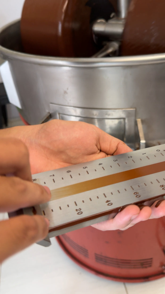 Hands checking ground cacao fineness against a micron gauge over the stone grinder