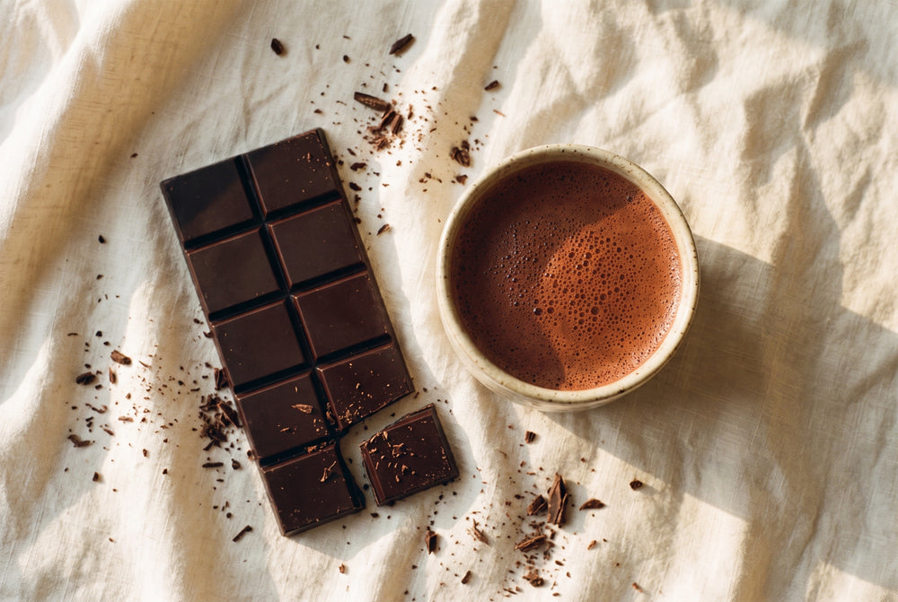 A cup of thick cacao beside the ten-piece NUDE bar on linen, shavings scattered around